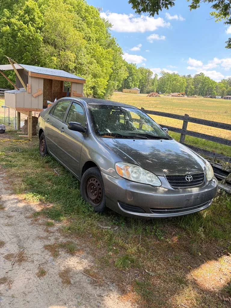 Older Toyota Corolla sitting unused beside a rural property before being sold as a junk car